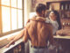 young men with bare torso is holding his beautiful girlfriend in arms while standing in kitchen at home
