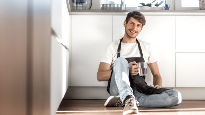 Young man with a glass of water sitting on the floor in the home kitchen.