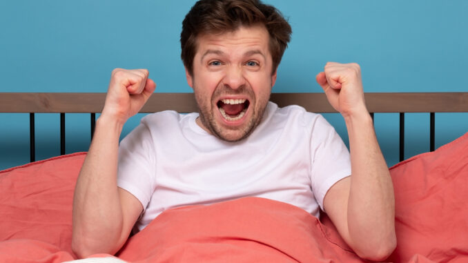 Caucasian positive man holding fists up resting at his bed at home