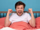 Caucasian positive man holding fists up resting at his bed at home
