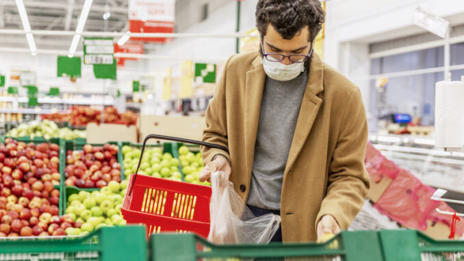 A young man in a medical mask is choosing fruits in a large supermarket.