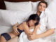 An image of a sweet young couple cuddling on a bed with white sheets and pillows