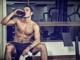Attractive athletic shirtless young man drinking protein shake from blender in gym while looking at camera