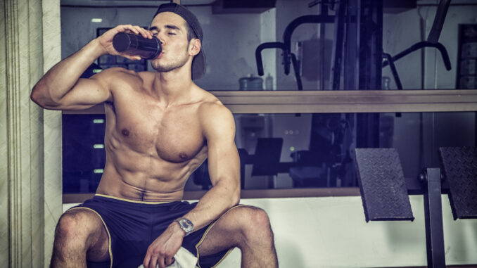 Attractive athletic shirtless young man drinking protein shake from blender in gym while looking at camera