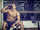 Attractive athletic shirtless young man drinking protein shake from blender in gym while looking at camera