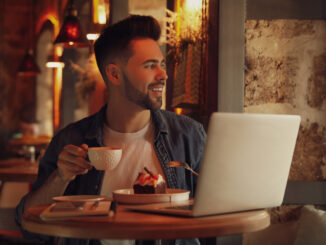 Young blogger with laptop eating dessert and drinking coffee at table in cafe