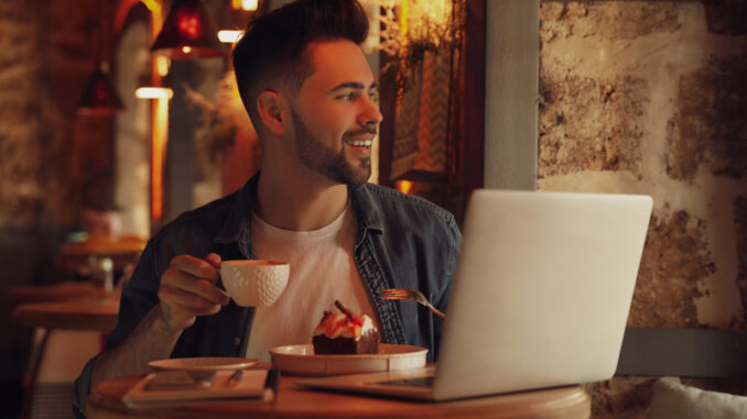 Young blogger with laptop eating dessert and drinking coffee at table in cafe