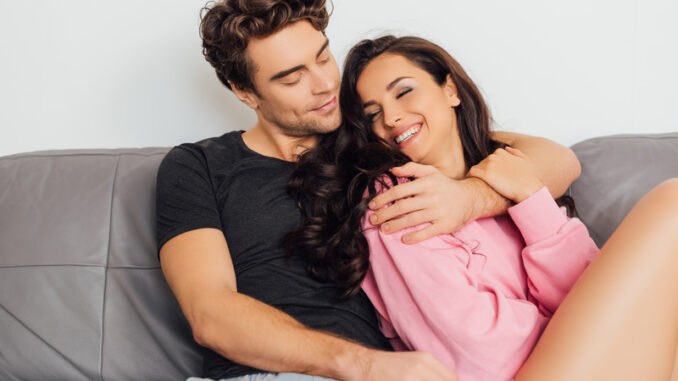 Selective focus of smiling woman touching boyfriend on sofa on grey