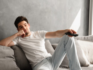 Photo of young bored man sitting on couch at home and changing TV channels with disinterest