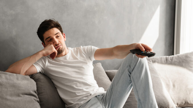 Photo of young bored man sitting on couch at home and changing TV channels with disinterest