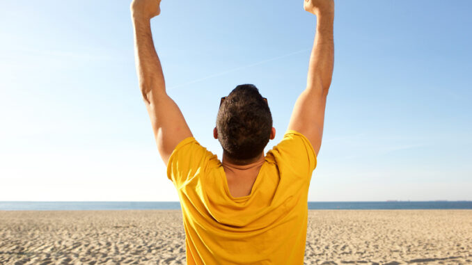 Portrait from back of man cheering with arms raised at the beach