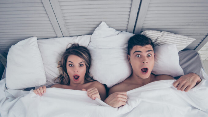 funny men and women lying in bed covered by sheets with wide opened eyes