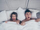 funny men and women lying in bed covered by sheets with wide opened eyes