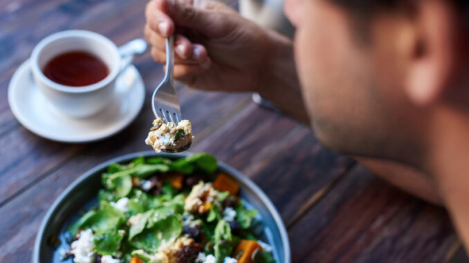 Young man sitting alone at a bistro table eating a delicious plate of mixed salad with a fork and drinking a cup of tea