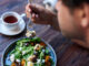 Young man sitting alone at a bistro table eating a delicious plate of mixed salad with a fork and drinking a cup of tea