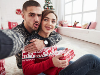Man taking selfie of him and his wife dressed in the Christmas clothes and sitting on the floor of decorative beautiful room.