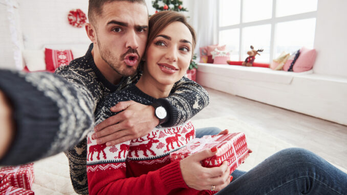 Man taking selfie of him and his wife dressed in the Christmas clothes and sitting on the floor of decorative beautiful room.