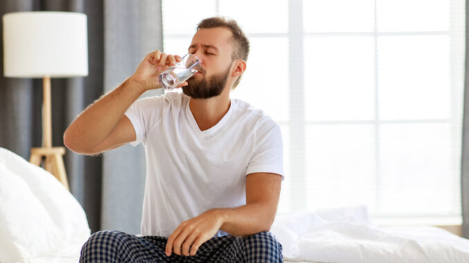 Young healthy man drinking water in morning at window