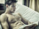 handsome young man laying shirtless on his bed next to window, reading a book