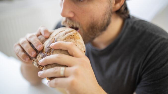 Surprised-looking man at white table calmly eating bread.