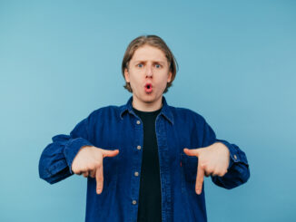Surprised young man in a shirt stands on a blue background and shows his hands down on the copy space
