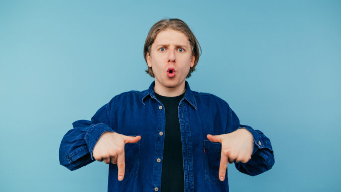 Surprised young man in a shirt stands on a blue background and shows his hands down on the copy space